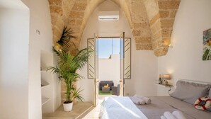 Un dormitorio con cama, una planta en maceta, una mesita de noche con lámpara y vistas a un balcón por una puerta abierta.
