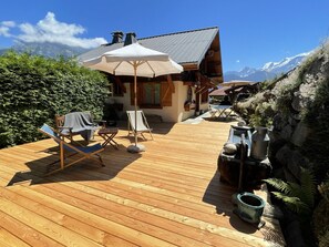 Un terrazzo in legno con mobili da esterno, un ombrellone grande e un edificio rustico con vista sulle montagne.