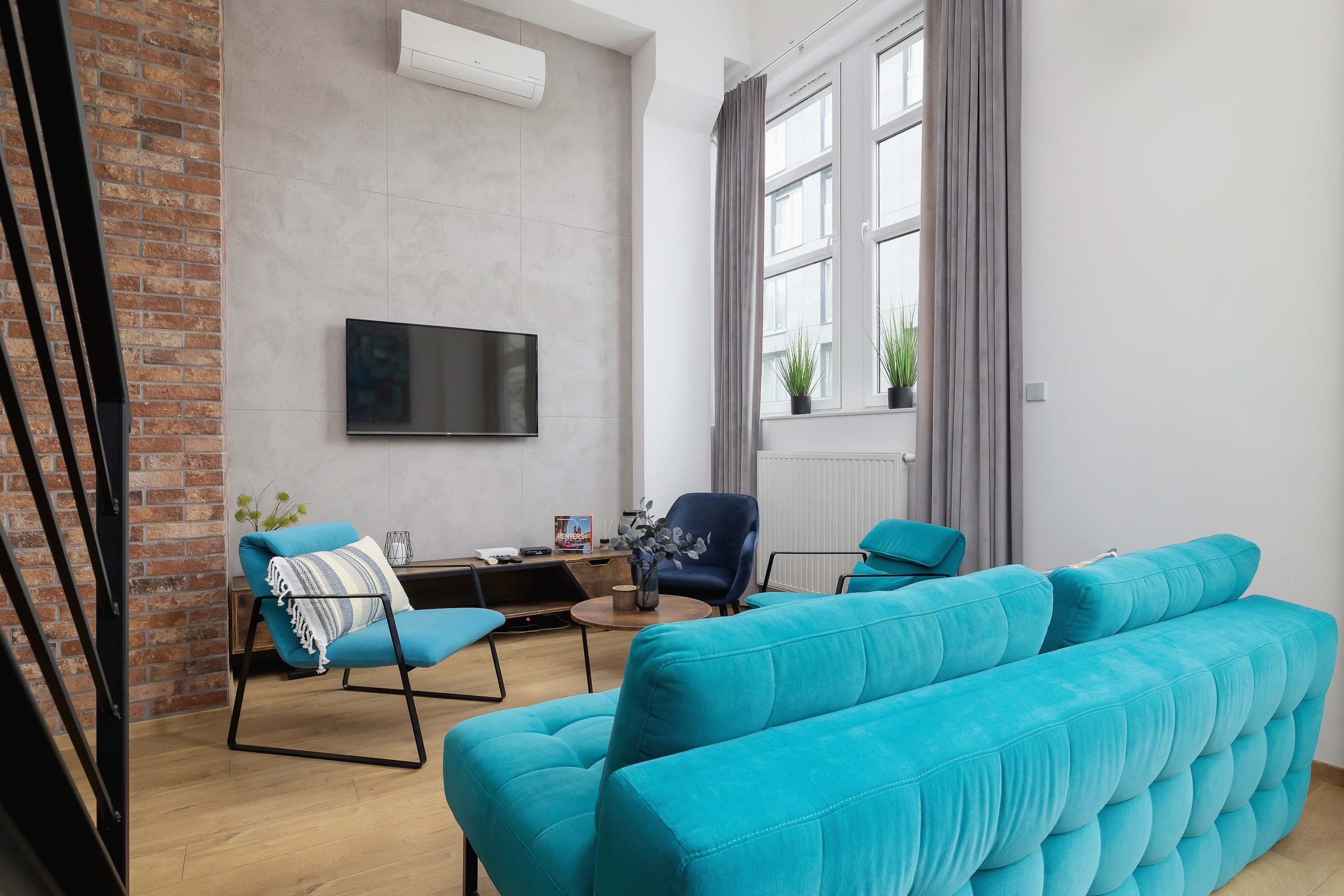 A modern living room with a large teal sofa, a blue armchair, and a flat-screen TV mounted on the wall.