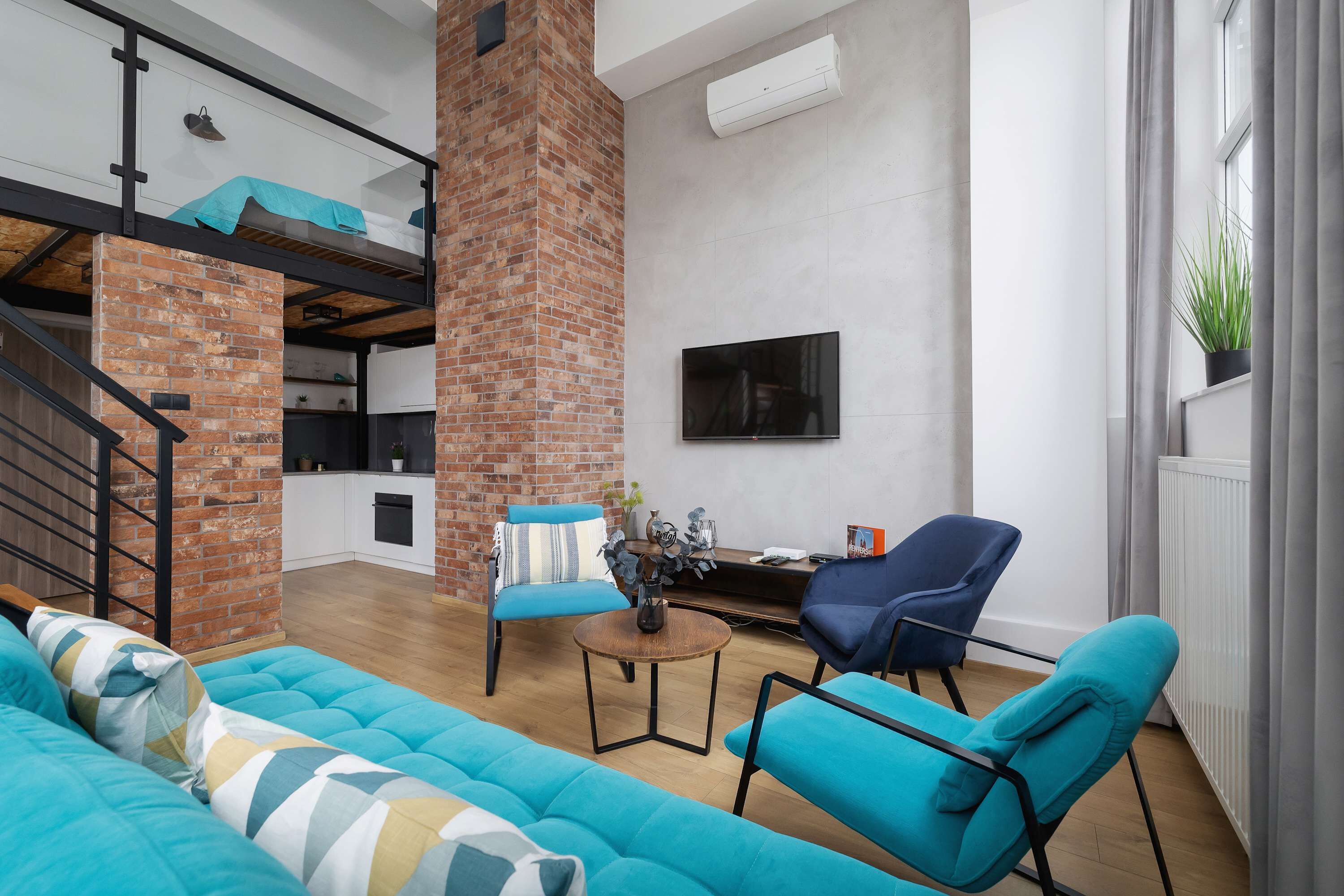 A modern living room with a blue sofa, a coffee table, and two blue armchairs. A brick column and a staircase are visible in the background.