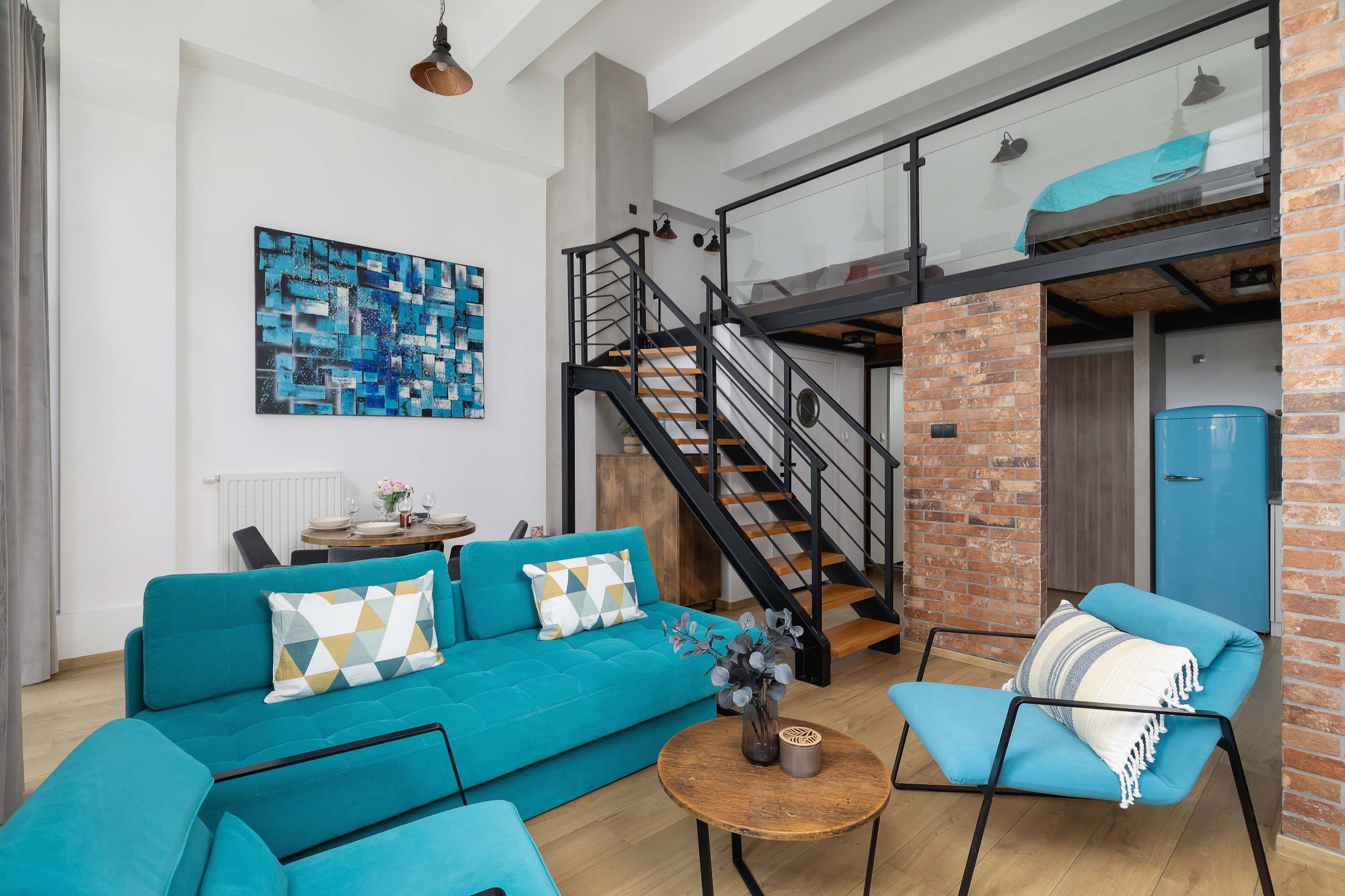 A modern living room with a teal sofa, a blue armchair, and a wooden coffee table. A staircase with a glass railing leads to a loft area.