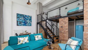 A modern living room with a teal sofa, a blue armchair, and a wooden coffee table. A staircase with a glass railing leads to a loft area. - Stylish Studio with Mezzanine | Air Conditioning | Close to the Center (Kraków)