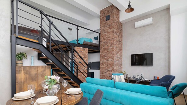 A modern living space with a staircase, a brick fireplace, a teal sofa, and a dining area with a wooden table set for four. - Stylish Studio with Mezzanine | Air Conditioning | Close to the Center (Kraków)