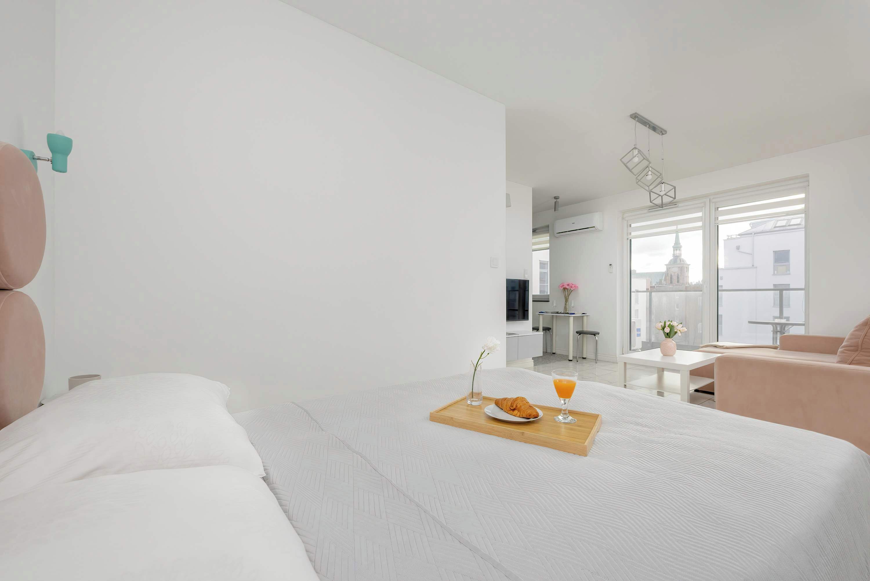 Una habitación de hotel moderna con una cama grande, una bandeja de desayuno con croissant y jugo de naranja, una mesita con flores, y vistas de la ciudad a través de amplias ventanas.