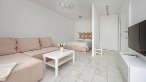 A modern, minimalist living room with a pink sofa, a white coffee table, a bed with a circular headboard, and a flat-screen TV.