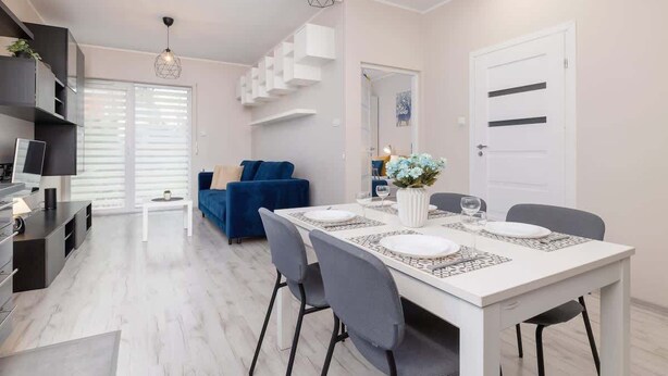 Una sala de estar moderna con un sofá azul, una mesa de comedor para cuatro personas y una cocina con armarios blancos y una encimera negra.