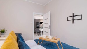 Habitación de hotel con cama, mesita de noche con bandeja que contiene tazas y una vela, y vista al área de la cocina a través de una puerta abierta.