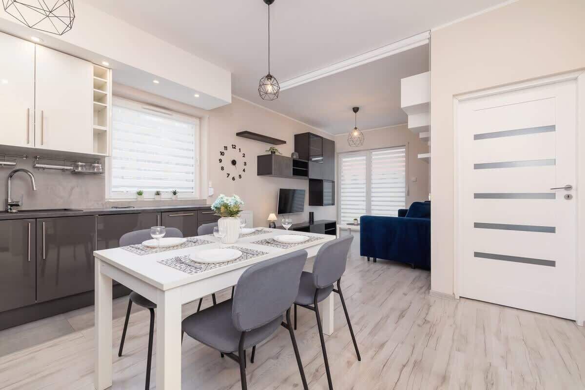 Ett modernt kök med en matplats, med ett vitt bord för fyra personer, grå stolar och en klocka på väggen.