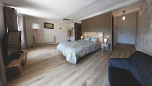 Um quarto espaçoso com uma cama grande, piso de madeira, sofá azul escuro, abajur e um guarda-roupa.