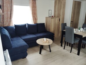A living room with a dark blue sofa, a round wooden coffee table, and a dining area with a wooden table and chairs. - Mobilhome PMR (Seignosse)
