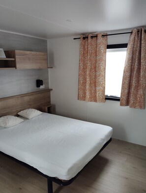 A single bed with a wooden headboard, a bedside table, and a window with patterned curtains. - Mobilhome PMR (Seignosse)
