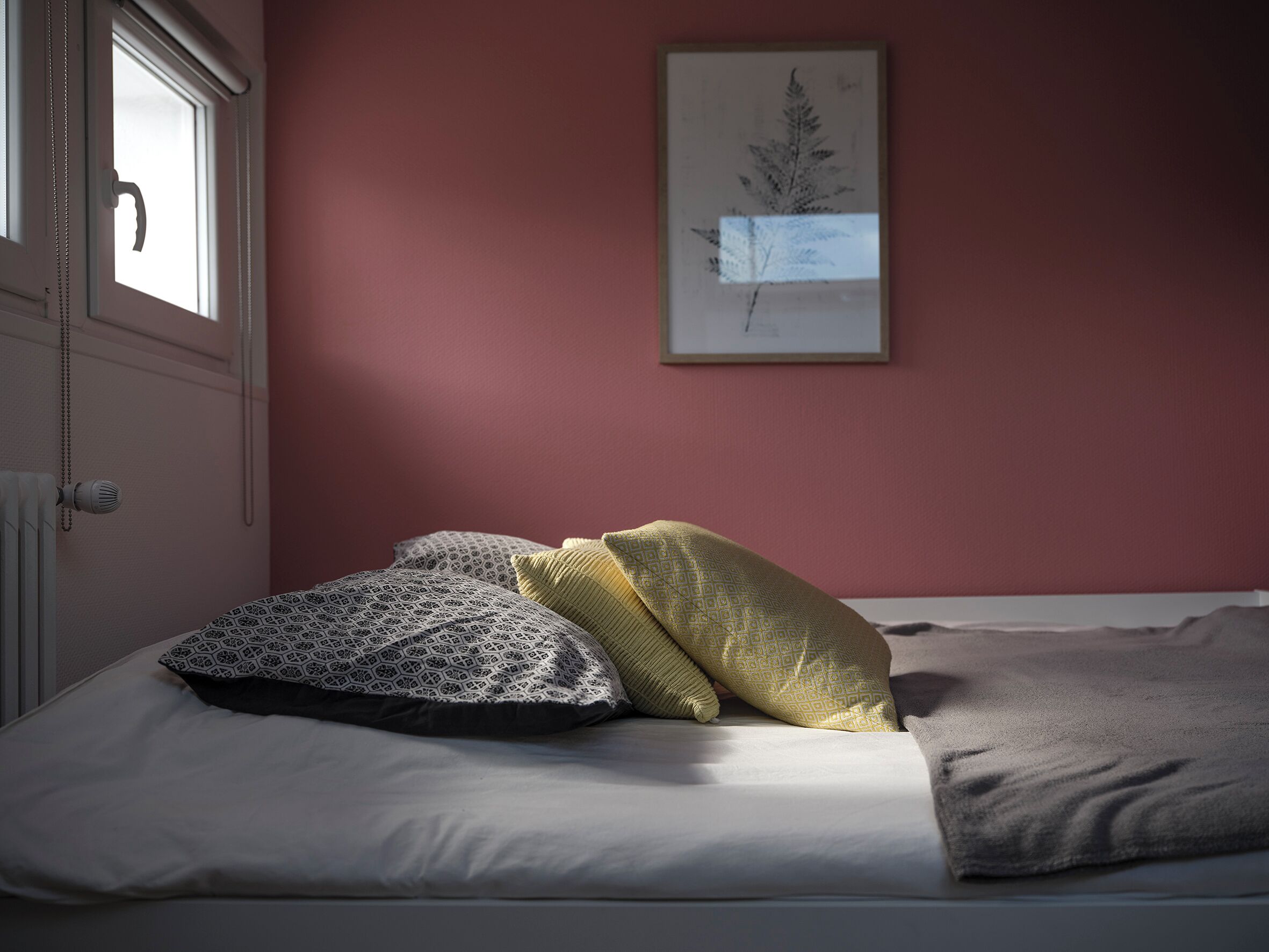 A bed with patterned pillows, a framed botanical print on the wall, and a window with a view.