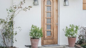A wooden door with a glass panel, flanked by two potted plants, set against a white wall with a textured finish.