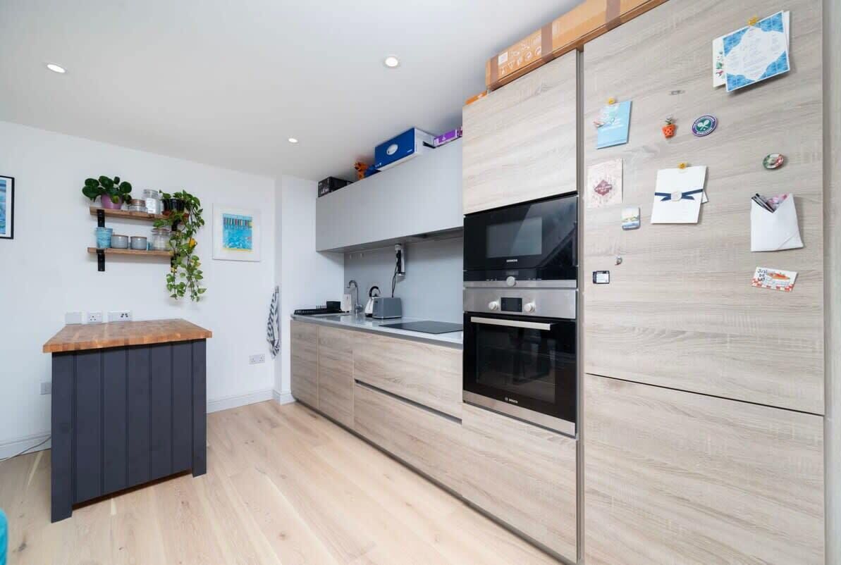 Una cucina moderna con mobili impiallacciati in legno, forno a incasso e microonde. Si vedono mensole con piante e accessori da cucina, e un piano di lavoro in legno.