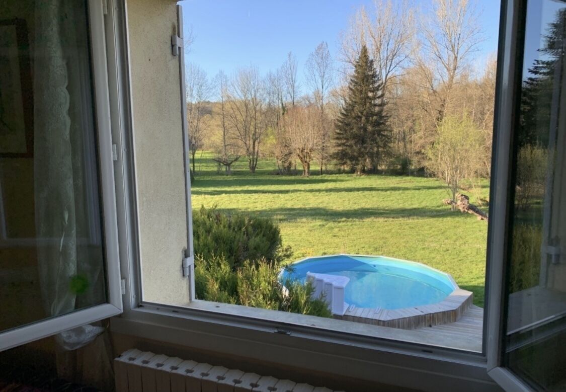 A view through a window of a backyard with a swimming pool, a grassy lawn, and trees.