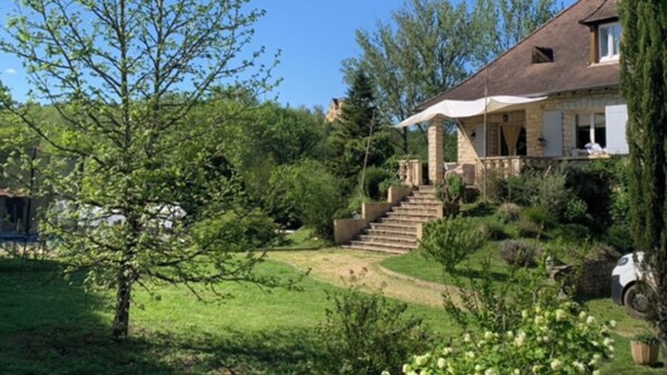 Une maison avec une façade en pierre, un porche abrité et un jardin avec une pelouse et des arbres.