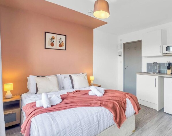 Ein Schlafzimmer mit Bett, Nachttischlampen, einem Bild an der Wand und einer Küchenzeile mit Spüle und Schränken.
