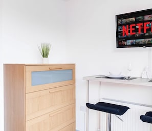 A modern kitchen with a wooden cabinet, a small plant, a bar area with stools, and a TV displaying Netflix content.