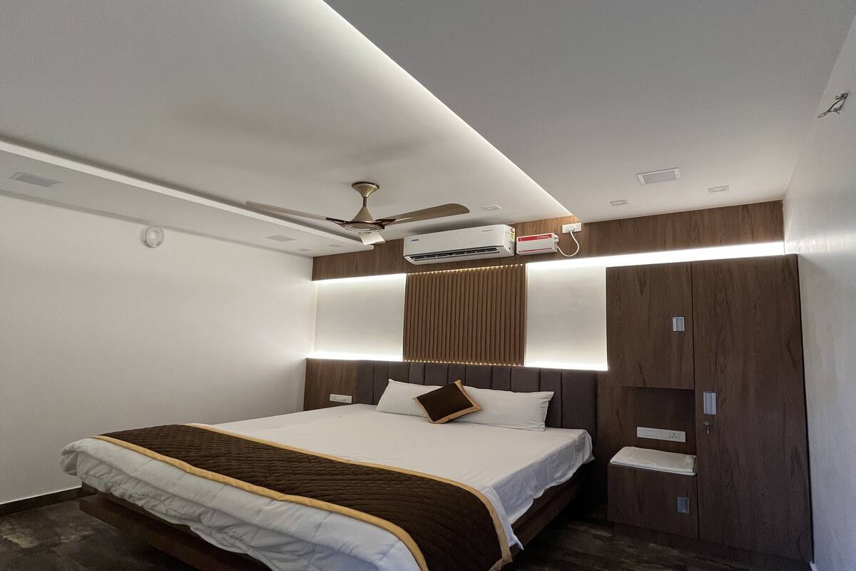 A modern hotel room with a large bed, built-in wardrobe, ceiling fan, and air conditioning unit.