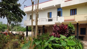 A two-story building with a flat roof, a balcony on the upper floor, and a garden with various plants in the foreground. - GP Village Resort (Melmalayanur)