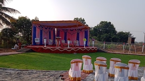 An outdoor stage with a red and blue backdrop, surrounded by a grassy area with white chairs with gold sashes. - GP Village Resort (Melmalayanur)