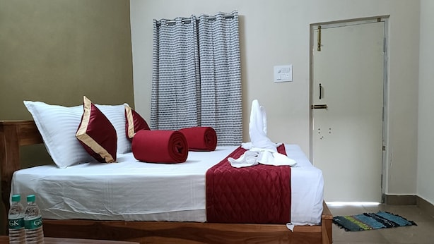 A neatly made bed with white linens, red and beige pillows, and a red blanket. There is a wooden nightstand with two water bottles. The room has a tiled floor and a door to the right. - GP Village Resort (Melmalayanur)