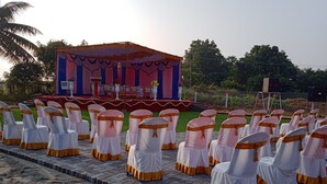 A stage with a backdrop, chairs with white covers and orange sashes, and a clear sky. - GP Village Resort (Melmalayanur)