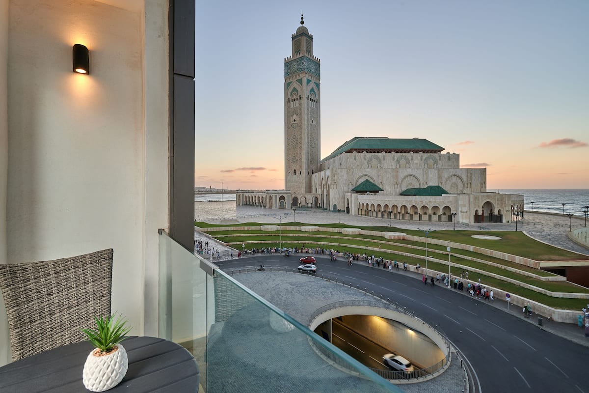 A view of a historic building with a tall tower and a green-roofed structure, a roundabout, and a body of water in the background.
