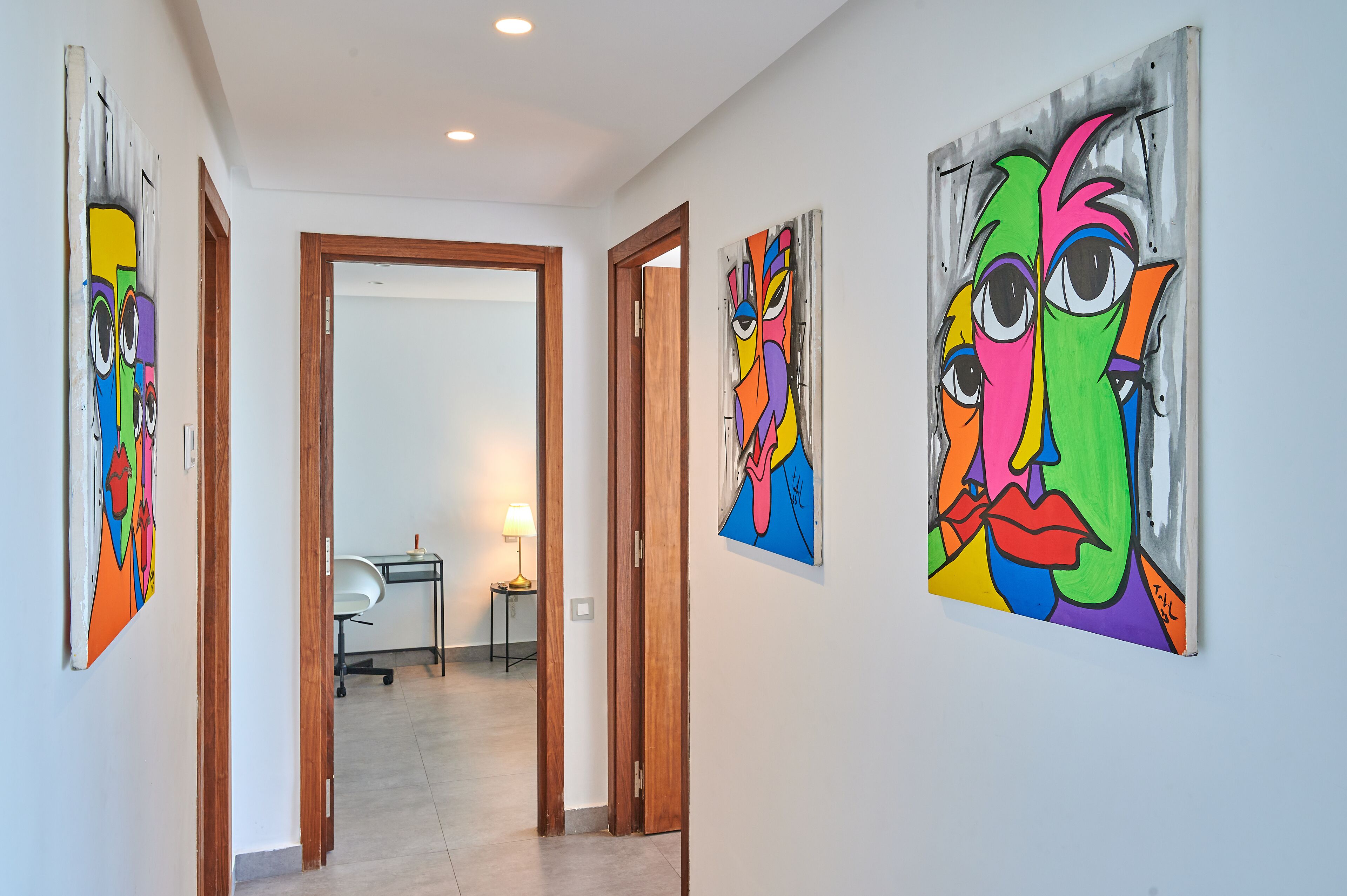 Un pasillo con tres cuadros abstractos y coloridos en las paredes, un marco de puerta de madera y un escritorio con una lámpara al fondo.