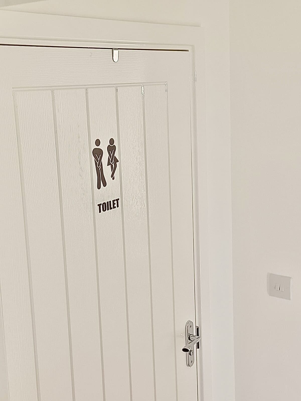Una puerta blanca con un letrero de baño que muestra dos figuras, una de pie y otra sentada, y la palabra TOILET debajo.