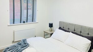 Uma cama bem arrumada com lençóis brancos, cabeceira cinzenta e mesa de cabeceira com abajur.