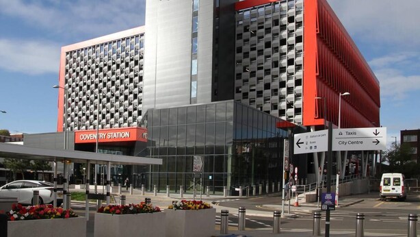 Um edifício moderno com um destaque para a placa da estação Coventry, grandes janelas e uma combinação de elementos arquitetônicos em vermelho e cinza.