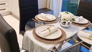 Uma área de refeições com mesa de vidro posta para quatro pessoas, pratos brancos, talheres dourados e um vaso com flores brancas.