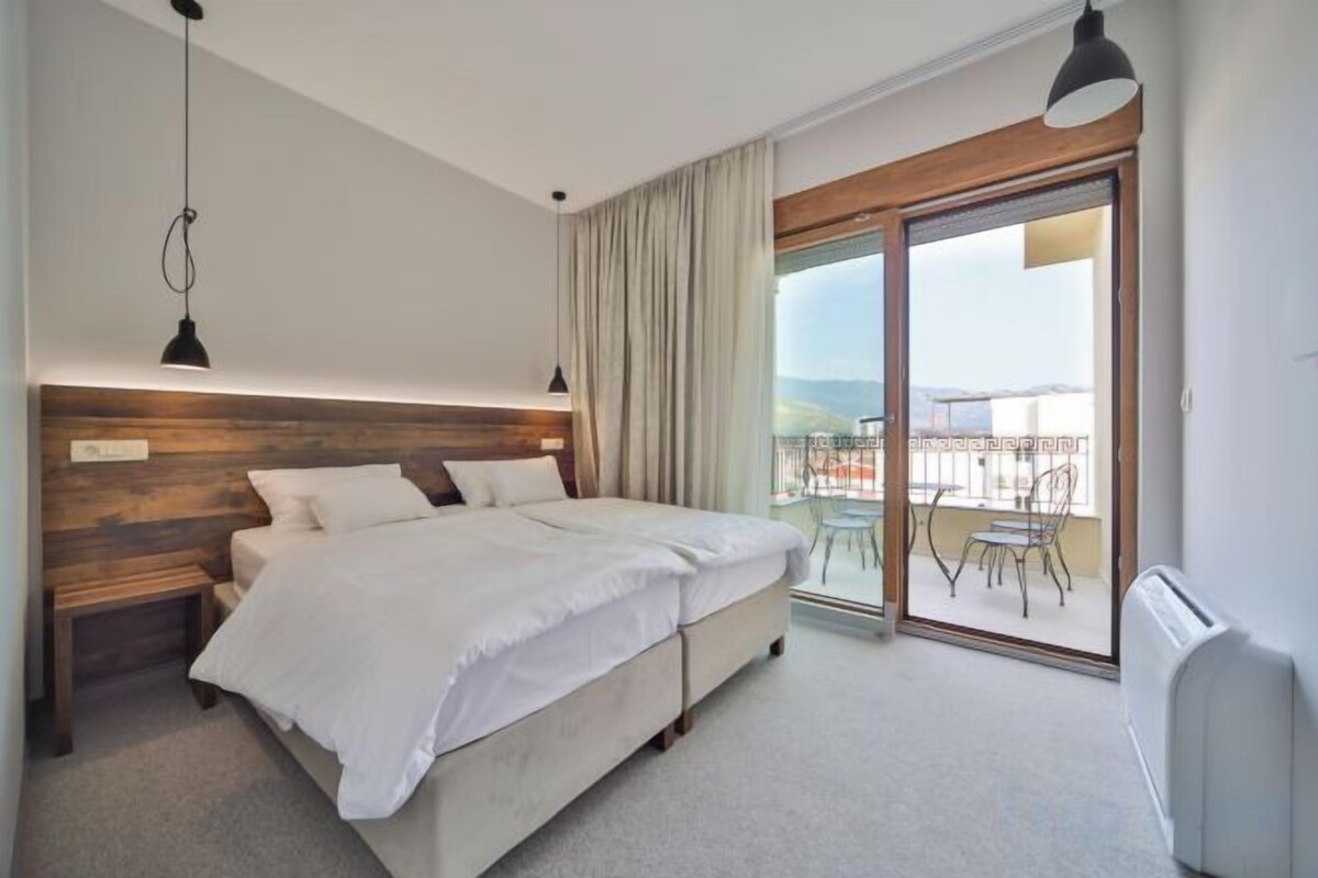 A bedroom with a large bed, a wooden headboard, a nightstand, and a view of a balcony with outdoor furniture.