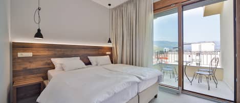A modern bedroom with a large bed, wooden headboard, and a view of a balcony with outdoor furniture.