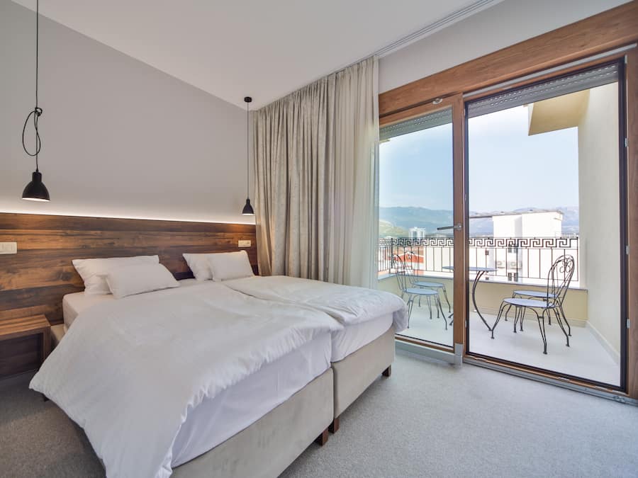 A modern bedroom with a large bed, wooden headboard, and a view of a balcony with outdoor furniture.