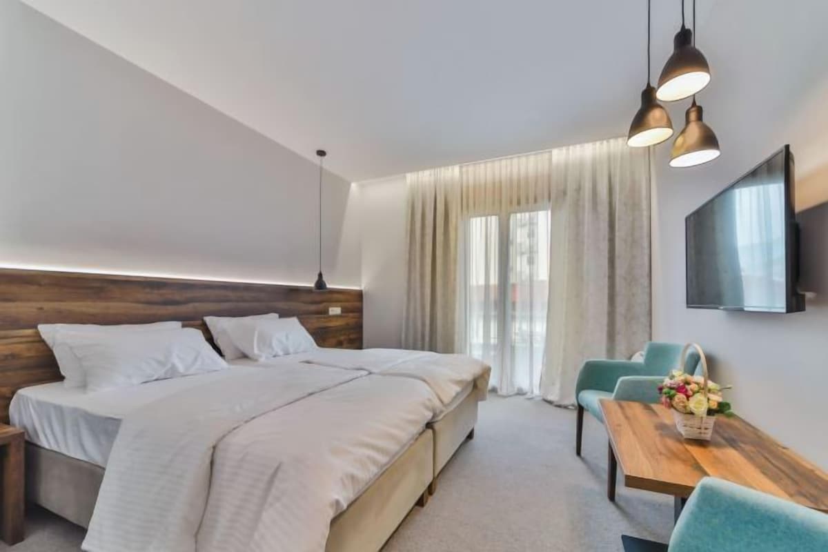 A modern hotel room with a large bed, a desk with a chair, a TV, and a window with curtains.