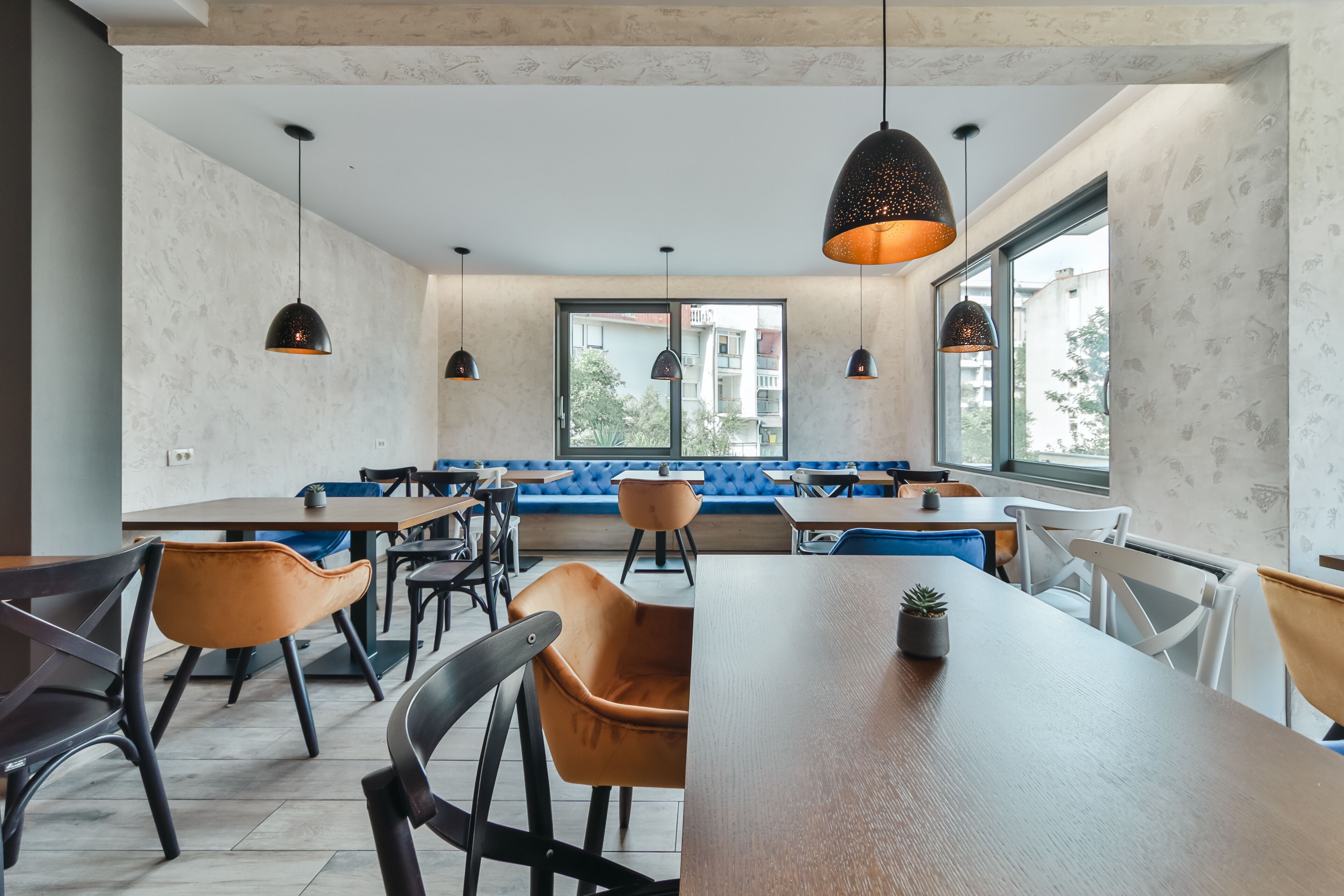 Ein moderner Essbereich mit Holztischen und -stühlen, blau gepolsterten Sitzgelegenheiten und Pendelleuchten.