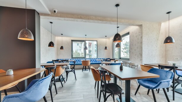 A modern dining area with wooden tables and blue chairs, large windows, and pendant lighting. - Hotel Vladimir (Budva)