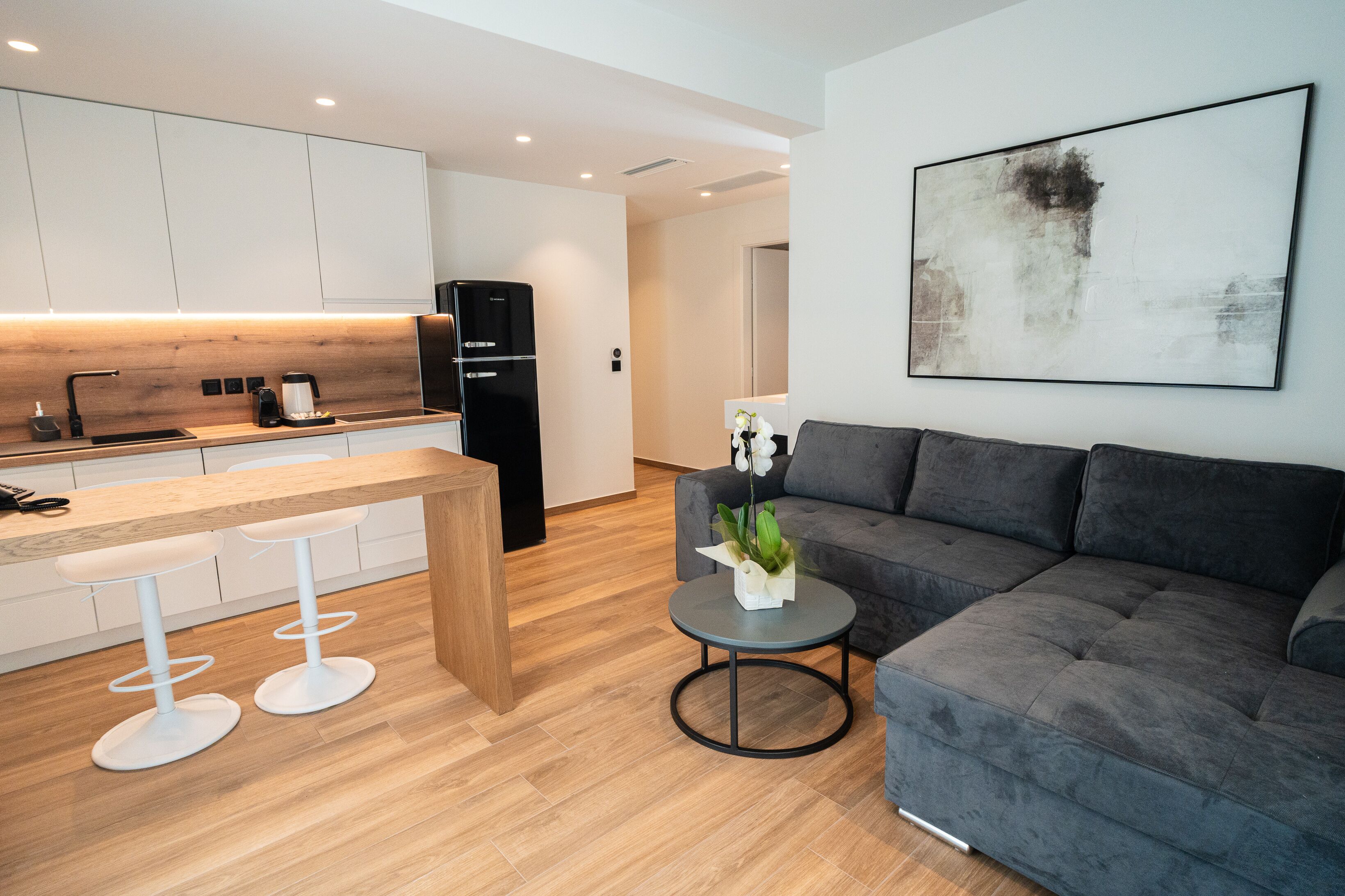 Et moderne køkken med hvide skabe, en træbordplade og to hvide barstole. Der er en grå sofa og et lille rundt bord med en plante i opholdsområdet.
