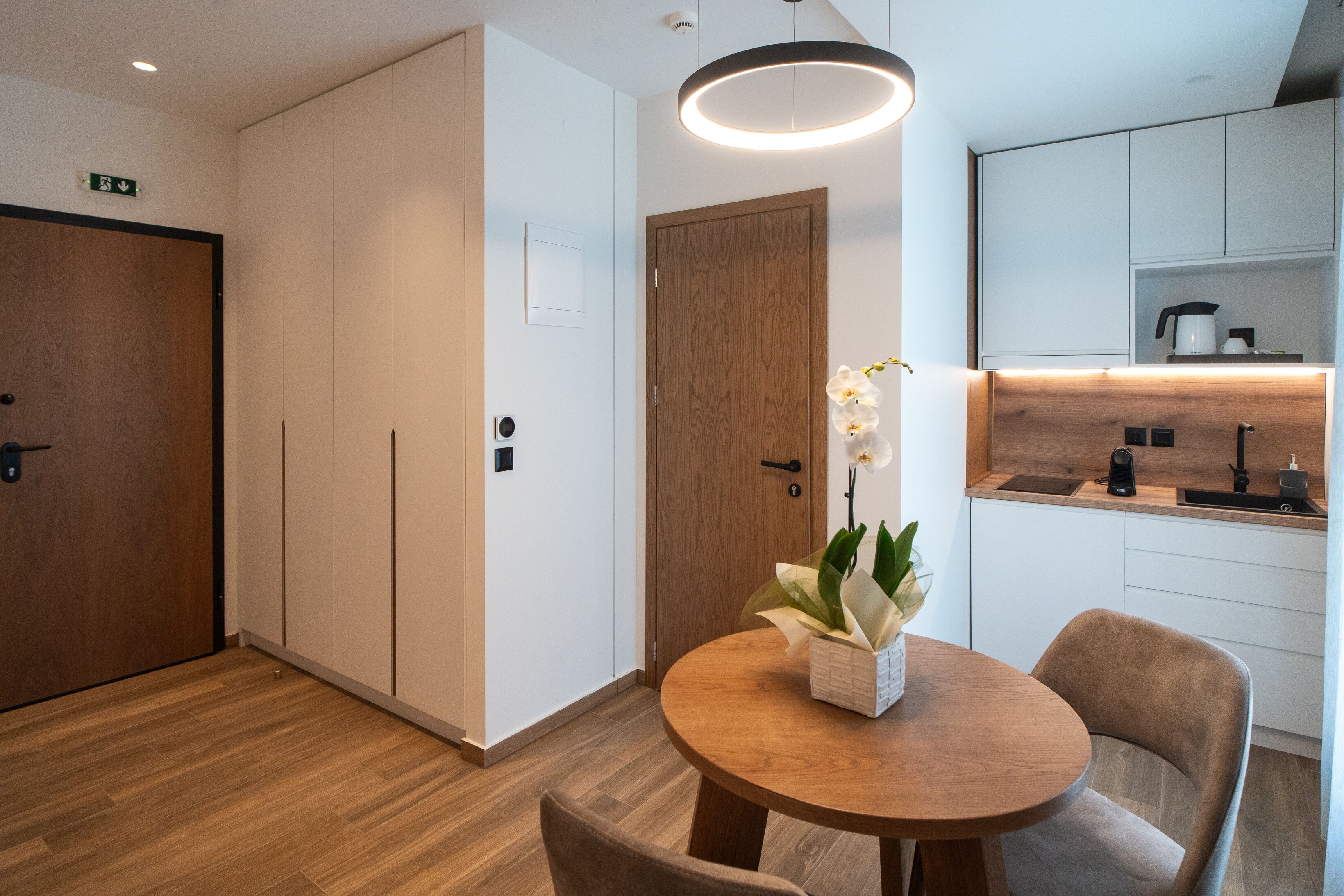 A modern hotel room with a wooden floor, a round dining table with chairs, a wooden door, and a kitchen area with white cabinets and a built-in oven.
