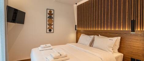 Une chambre d’hôtel moderne avec une tête de lit en bois, un téléviseur à écran plat fixé au mur et des serviettes soigneusement pliées sur le lit.