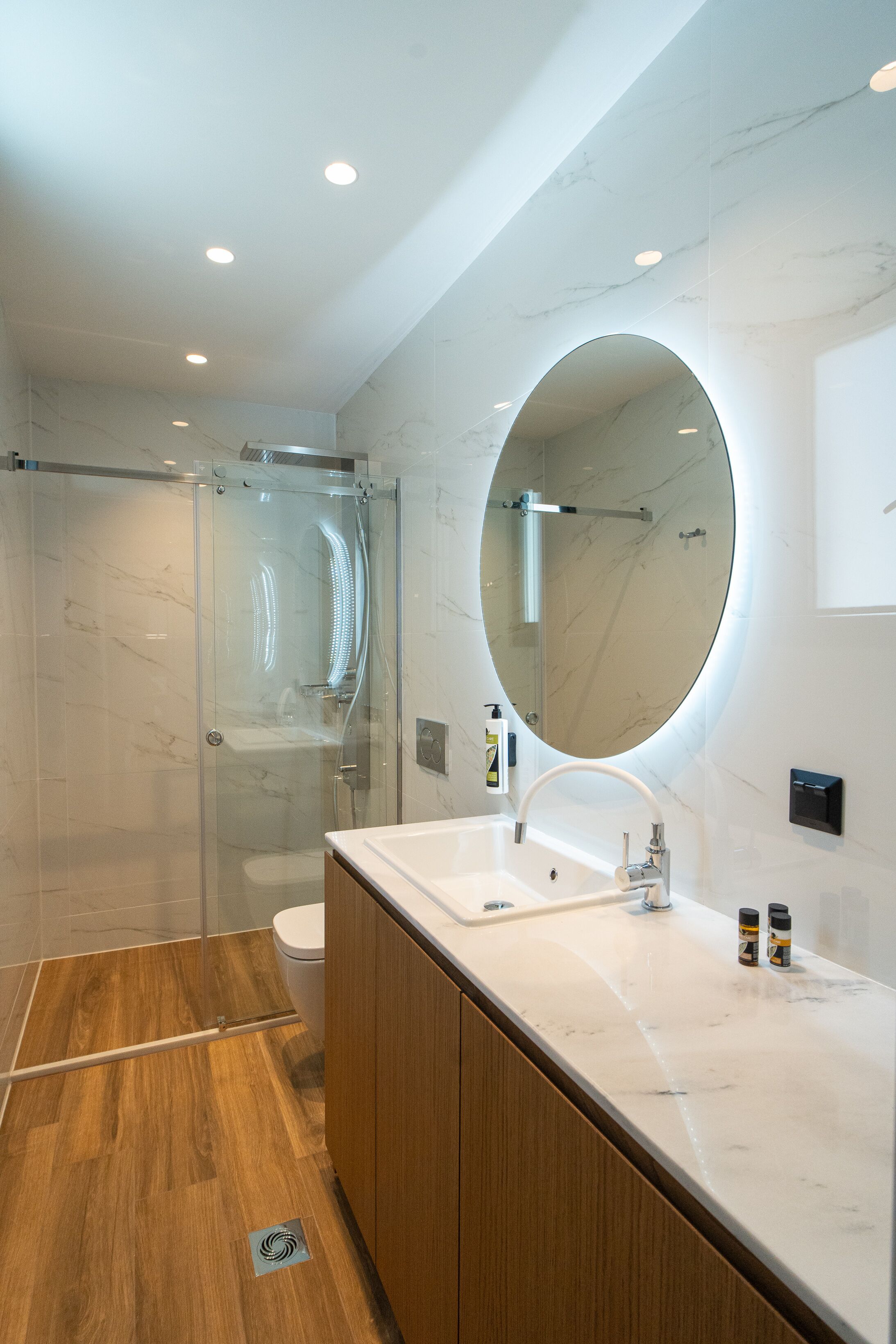 A modern bathroom with a glass-enclosed shower, a large oval mirror, a white sink, and wooden cabinetry.