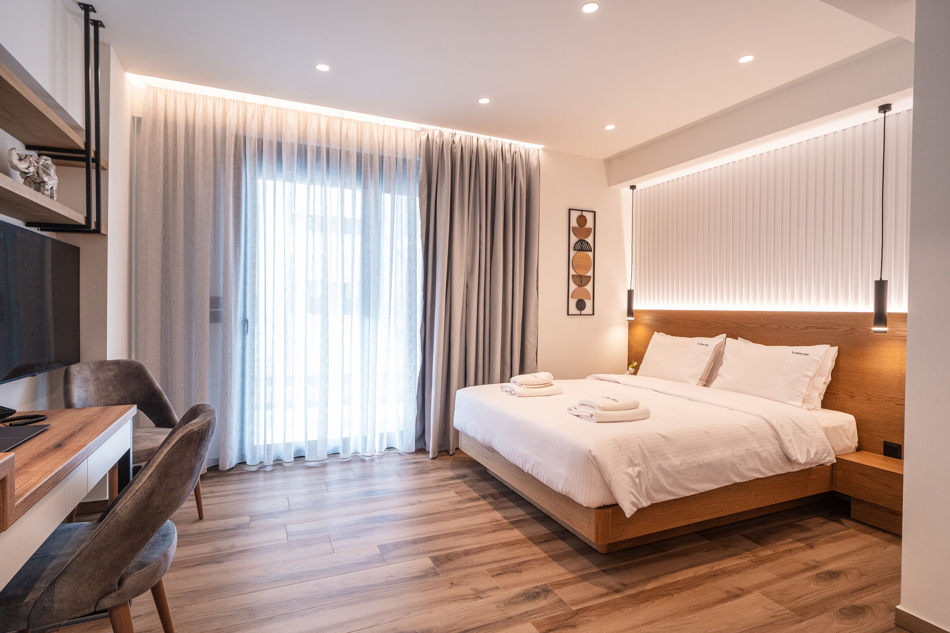 A modern hotel room with a wooden bed, a desk with a chair, and a large window with curtains.