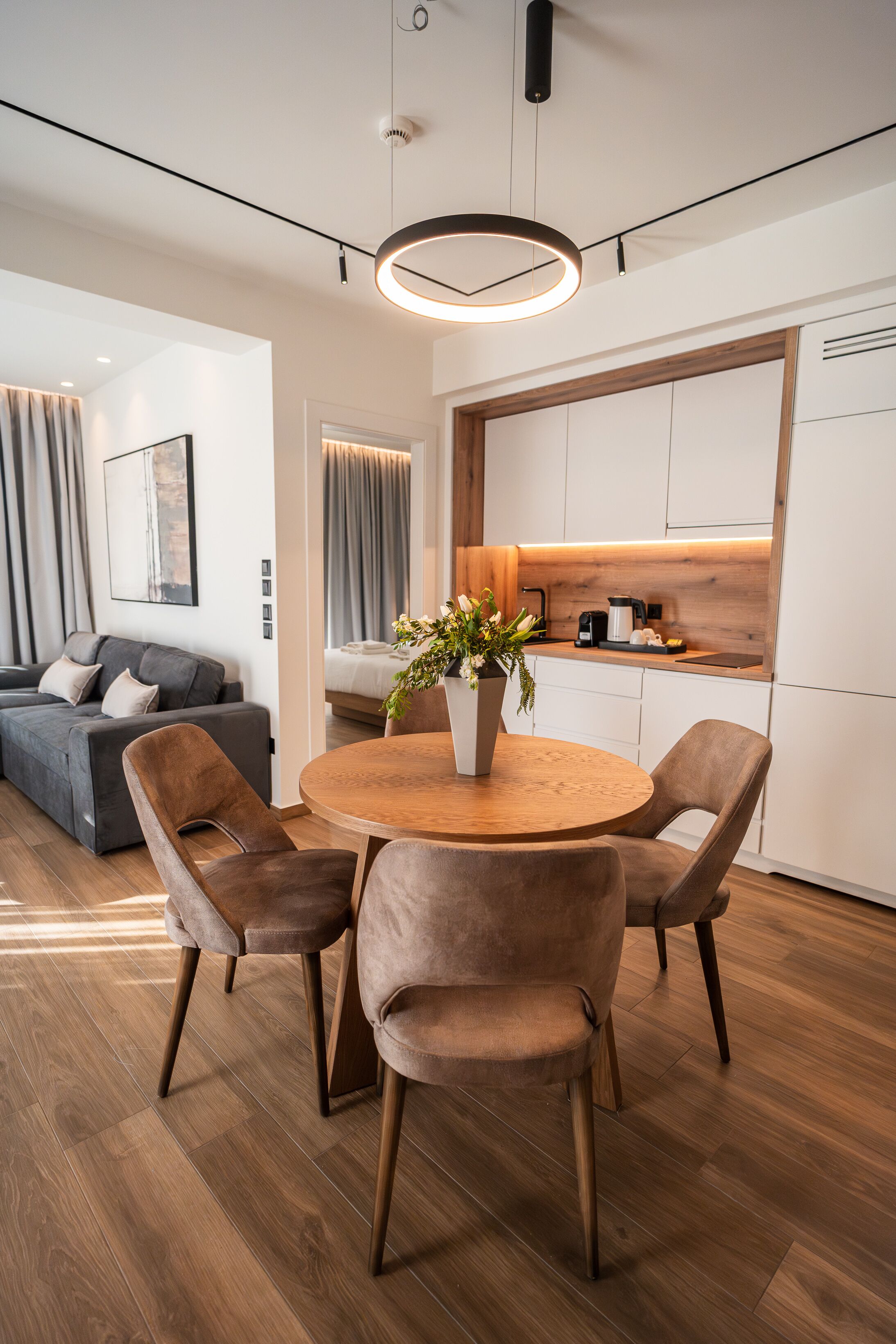 Una sala de estar moderna con una mesa redonda de madera, cuatro sillas acolchadas, un sofá gris y una zona de cocina con armarios blancos y encimera de madera.