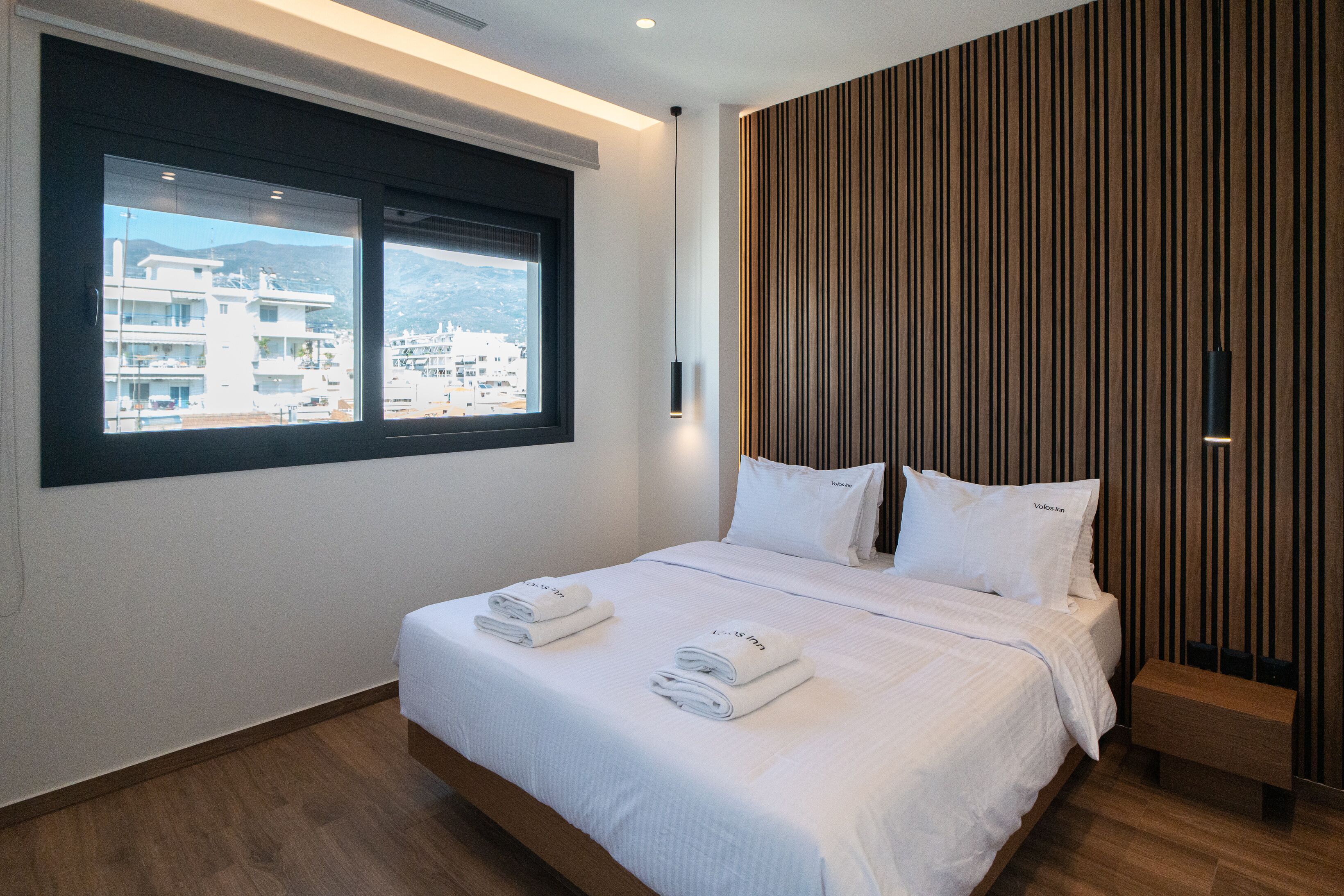 A modern hotel room with a large bed, two windows, a wooden panel wall, and a view of buildings outside.