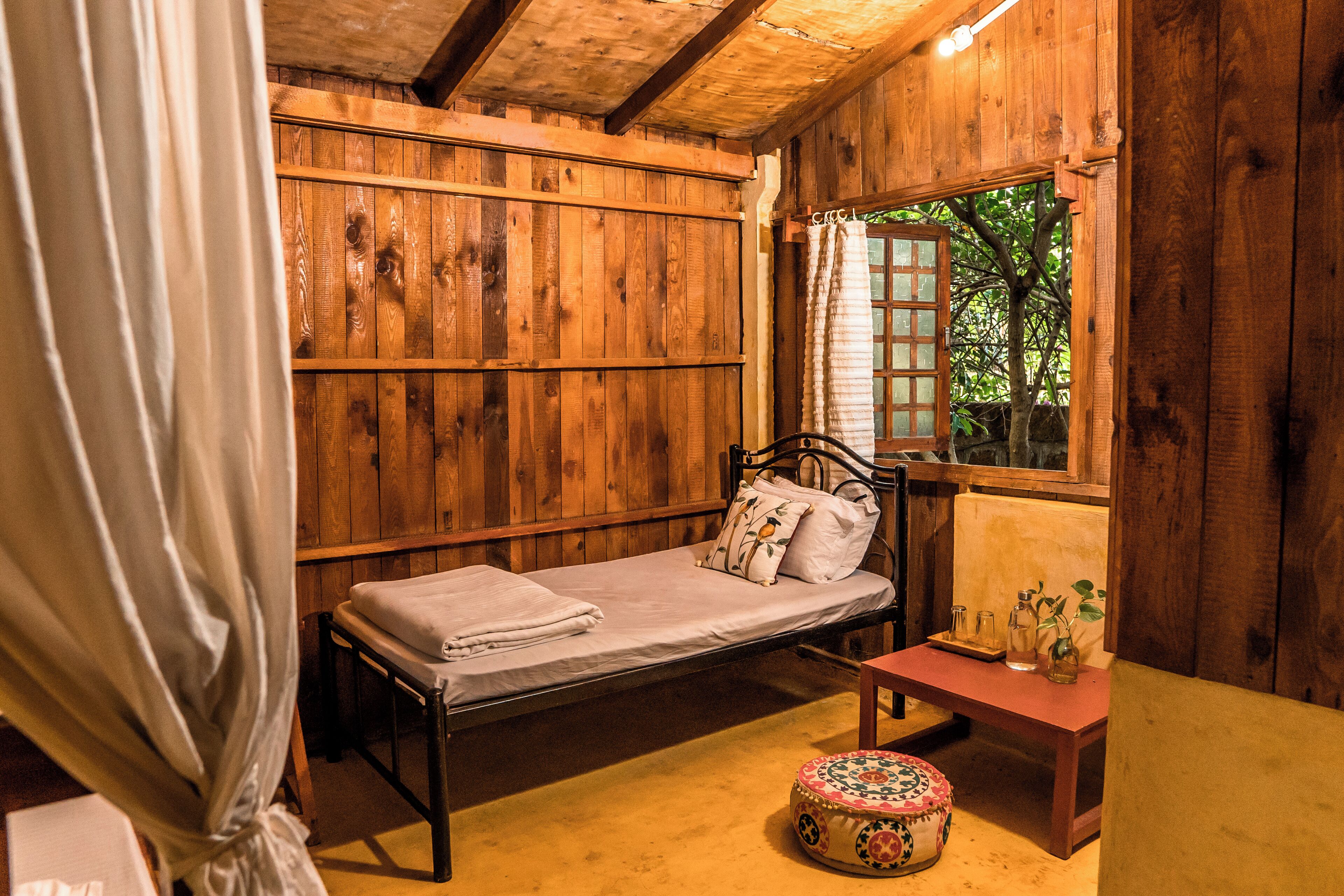 A wooden room with a single bed, a small table, and a window with curtains.