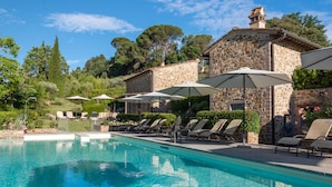 A swimming pool with lounge chairs and umbrellas, surrounded by stone buildings and greenery. - Tenuta Guardastelle (San Gimignano)