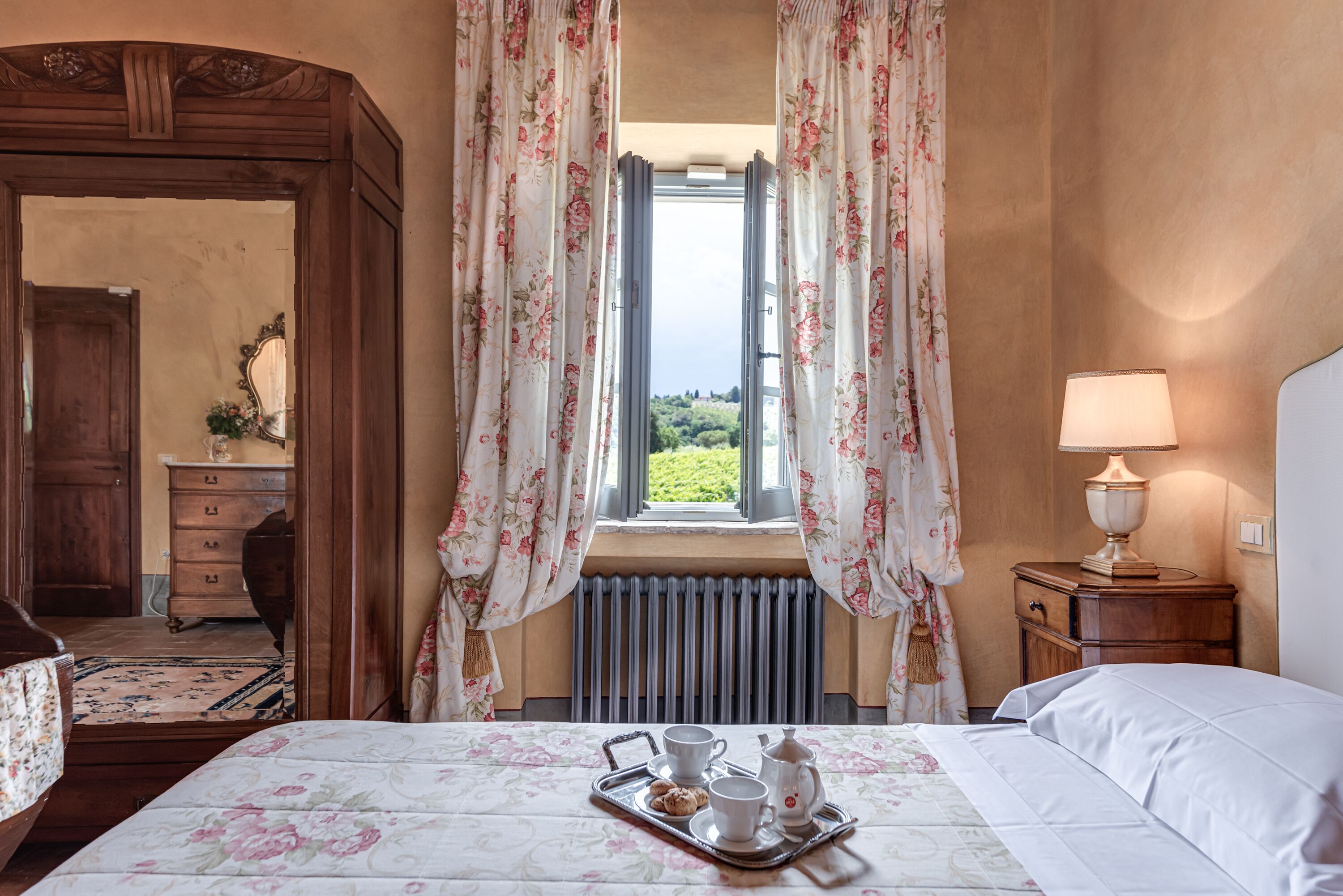 A bedroom with a bed, a nightstand with a lamp, a window with curtains, a radiator, and a tray with tea and cookies.