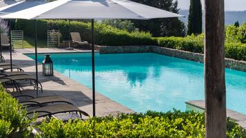 Una piscina con sillas de descanso y sombrillas, rodeada de vegetación y muros de piedra.